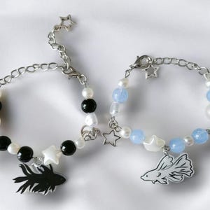 K&ouml;nnte beinhalten: Zwei Armb&auml;nder mit Perlen und silbernen Kettenakzenten. Ein Armband hat schwarze und wei&szlig;e Perlen mit einem schwarzen Fischanh&auml;nger. Das andere Armband hat blaue und wei&szlig;e Perlen mit einem wei&szlig;en Fischanh&auml;nger. Beide haben Sternanh&auml;nger.
