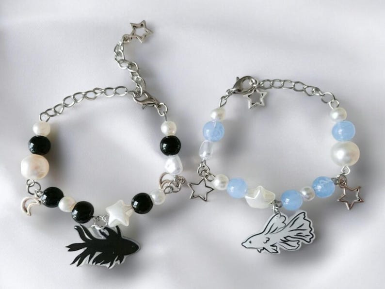 K&ouml;nnte beinhalten: Zwei Armb&auml;nder mit Perlen und silbernen Kettenakzenten. Ein Armband hat schwarze und wei&szlig;e Perlen mit einem schwarzen Fischanh&auml;nger. Das andere Armband hat blaue und wei&szlig;e Perlen mit einem wei&szlig;en Fischanh&auml;nger. Beide haben Sternanh&auml;nger.