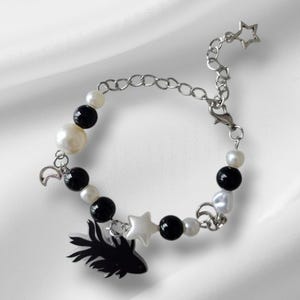 K&ouml;nnte beinhalten: Ein Armband mit schwarzen und wei&szlig;en Perlen, einer silbernen Kette und Anh&auml;ngern. Zu den Anh&auml;ngern geh&ouml;ren ein schwarzer Fisch, ein wei&szlig;er Stern und ein silberner Mond. Das Armband liegt auf wei&szlig;em Hintergrund.