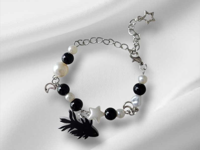 K&ouml;nnte beinhalten: Ein Armband mit schwarzen und wei&szlig;en Perlen, einer silbernen Kette und Anh&auml;ngern. Zu den Anh&auml;ngern geh&ouml;ren ein schwarzer Fisch, ein wei&szlig;er Stern und ein silberner Mond. Das Armband liegt auf wei&szlig;em Hintergrund.