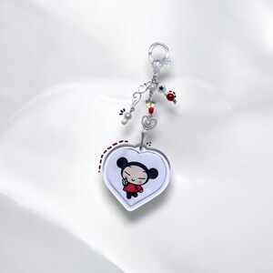K&ouml;nnte beinhalten: Ein wei&szlig;er herzf&ouml;rmiger Schl&uuml;sselanh&auml;nger mit einem Cartoon-Bild von Pucca, einer Figur mit schwarzen Haaren und einem roten Kleid, die ein Peace-Zeichen macht. Der Schl&uuml;sselanh&auml;nger hat einen silbernen Anh&auml;nger mit wei&szlig;en und roten Perlen.