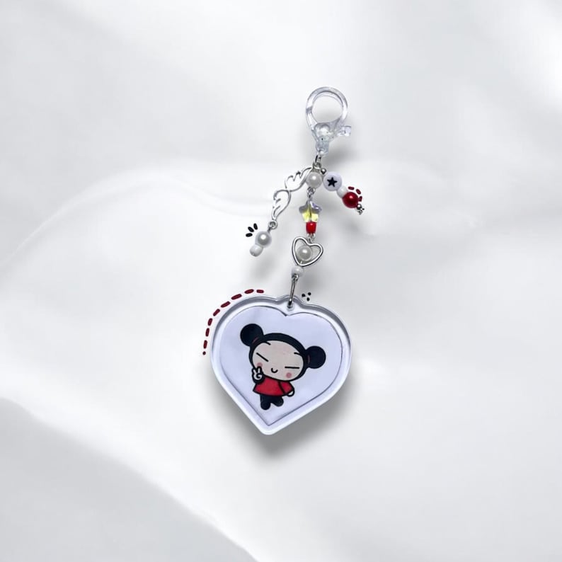 K&ouml;nnte beinhalten: Ein wei&szlig;er herzf&ouml;rmiger Schl&uuml;sselanh&auml;nger mit einem Cartoon-Bild von Pucca, einer Figur mit schwarzen Haaren und einem roten Kleid, die ein Peace-Zeichen macht. Der Schl&uuml;sselanh&auml;nger hat einen silbernen Anh&auml;nger mit wei&szlig;en und roten Perlen.