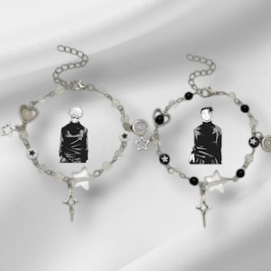Op de afbeelding: Twee zilveren armbanden met bedels. De ene armband heeft witte kralen en bedels, waaronder een hart, sterren en een personage uit een anime. De andere armband heeft zwarte kralen en bedels, waaronder een hart, sterren en een personage uit een anime.