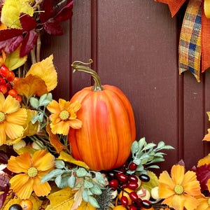 K&ouml;nnte beinhalten: Ein dekorativer Herbstkranz mit einem zentralen orangefarbenen K&uuml;rbis, gelben und burgunderroten Kunstbl&auml;ttern, orangefarbenen Kosmeenbl&uuml;ten sowie Trauben aus roten und gelben Beeren. Ein kariertes Band in Orange, Gelb und Blau ziert die rechte Seite.