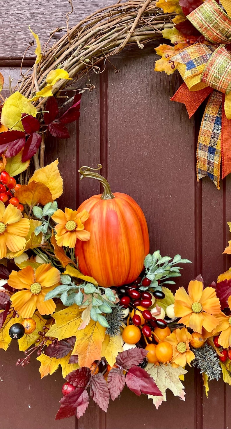 K&ouml;nnte beinhalten: Ein dekorativer Herbstkranz mit einem zentralen orangefarbenen K&uuml;rbis, gelben und burgunderroten Kunstbl&auml;ttern, orangefarbenen Kosmeenbl&uuml;ten sowie Trauben aus roten und gelben Beeren. Ein kariertes Band in Orange, Gelb und Blau ziert die rechte Seite.