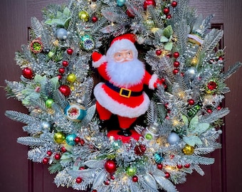 Könnte beinhalten: Ein festlicher Weihnachtskranz aus silberfarbenen künstlichen Tannenzweigen, geschmückt mit kleinen roten, grünen, blauen und silbernen Kugeln, roten Beerenclustern und warmweißen Lichtern, in der Mitte eine Weihnachtsmannfigur im roten Anzug.