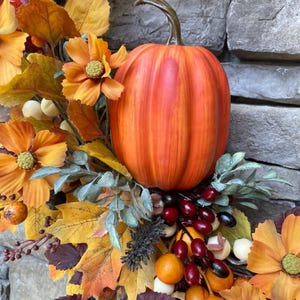 K&ouml;nnte beinhalten: Ein dekorativer Herbstkranz mit einem zentralen orangefarbenen K&uuml;rbis mit geschwungenem Stiel, umgeben von gelben und orangefarbenen Blumen, gold- und burgunderfarbenen Ahornbl&auml;ttern sowie kleinen roten und wei&szlig;en Beeren.