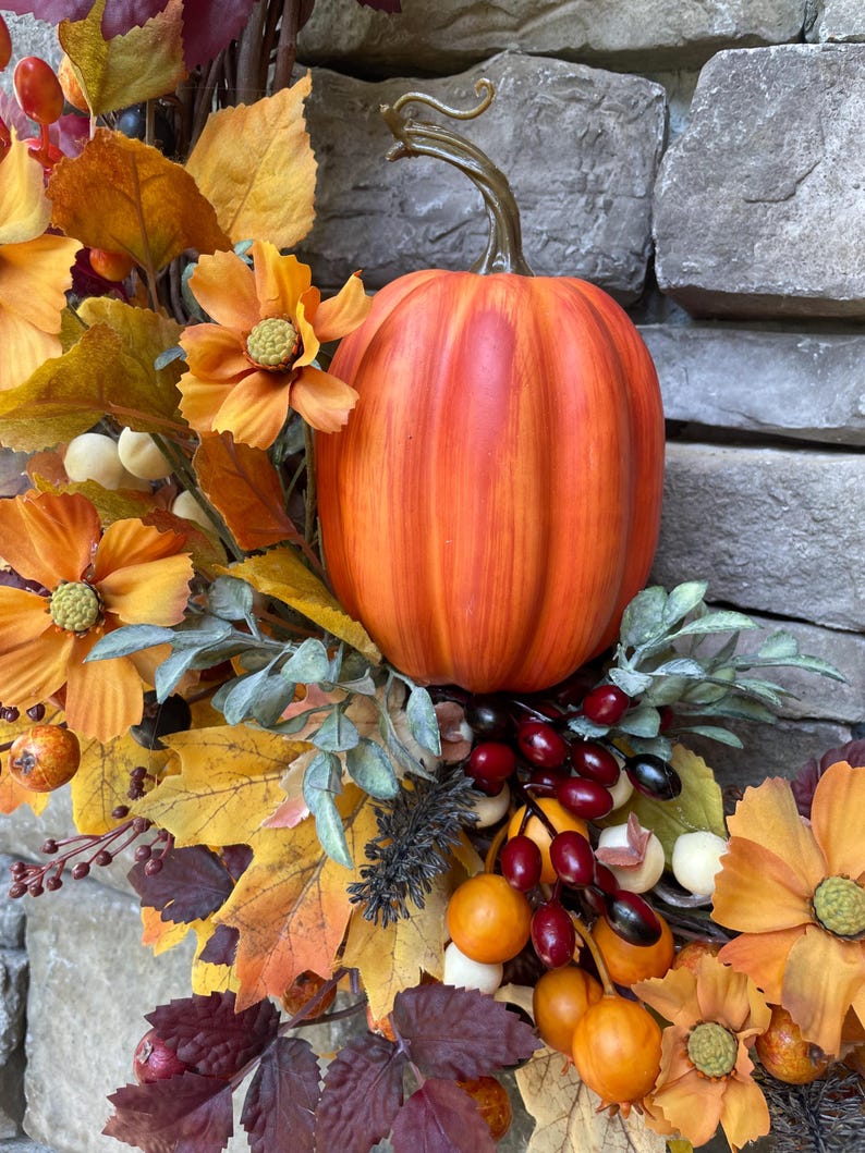 K&ouml;nnte beinhalten: Ein dekorativer Herbstkranz mit einem zentralen orangefarbenen K&uuml;rbis mit geschwungenem Stiel, umgeben von gelben und orangefarbenen Blumen, gold- und burgunderfarbenen Ahornbl&auml;ttern sowie kleinen roten und wei&szlig;en Beeren.