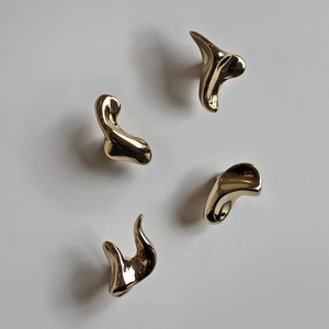 Può includere: Quattro ganci in metallo dorato astratto, ognuno con una forma organica unica. I ganci sono montati su una parete bianca.