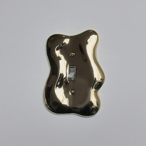 Puede incluir: Una cubierta de interruptor de luz de color latón con una forma orgánica y fluida. La cubierta tiene un acabado brillante y un interruptor basculante estándar. El diseño es moderno y escultural, añadiendo un toque artístico a cualquier habitación.