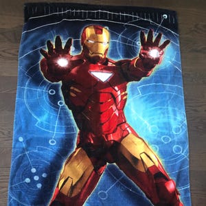 May include: A blue beach towel with a red and gold Iron Man character in a superhero pose. The towel has the text "IRON MAN 2" in gold letters and a small Marvel logo in the bottom left corner.