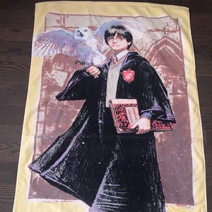 May include: A yellow beach towel with a cartoon image of Harry Potter holding a book and an owl. The towel has the text "Harry Potter" printed on it.