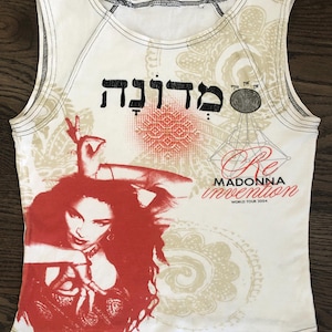 May include: White sleeveless shirt featuring a red and black graphic of a woman with long hair. The shirt includes text in Hebrew and the words "Re Invention Madonna World Tour 2004". The shirt has a black stitched neckline.