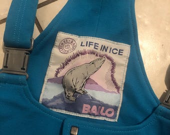 May include: Bright turquoise bib overalls with adjustable straps and metal hardware. A patch on the chest features a polar bear illustration with the text "LIFE IN ICE" and "BAILO".