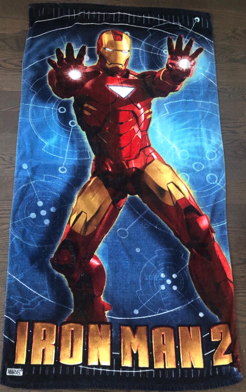 May include: A blue beach towel with a red and gold Iron Man character in a superhero pose. The towel has the text "IRON MAN 2" in gold letters and a small Marvel logo in the bottom left corner.