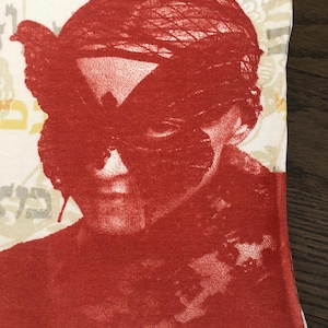 May include: Close-up of a white t-shirt with a red graphic print. The print features a face with a bird-like mask. The background has a floral pattern and Hebrew text. The shirt has a black trim.