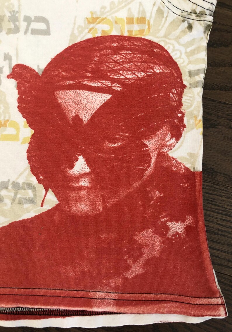 May include: Close-up of a white t-shirt with a red graphic print. The print features a face with a bird-like mask. The background has a floral pattern and Hebrew text. The shirt has a black trim.