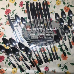 May include: A set of vintage silverware, including knives, forks, and spoons, laid out on a floral tablecloth. The silverware is made of stainless steel and has a polished finish. The text "1932 LADY HAMILTON FLATWARE" is visible in the image.