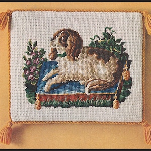 May include: A white needlepoint pillow with a brown and white dog lying on a blue cushion. The dog is surrounded by green foliage and pink flowers. The pillow has a gold tassel on each corner.