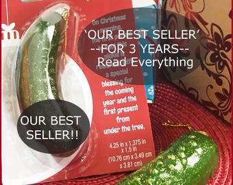May include: A green and gold glitter Christmas pickle ornament with a gold string for hanging. The ornament is packaged in a clear plastic bag with a red and white label that reads "The Tradition of the Christmas Pickle" and "Our Best Seller - For 3 Years - Read Everything". The label also includes the dimensions of the ornament: 4.25 inches x 1.375 inches x 1.5 inches (10.76 cm x 3.49 cm x 3.81 cm).