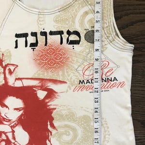 May include: White tank top with a red and beige graphic print. The design includes Hebrew text, a portrait, and the words "Re-Invention" and "Madonna". The tank top measures approximately 21 inches in length.