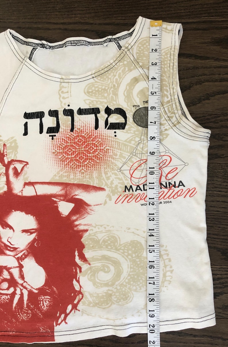 May include: White tank top with a red and beige graphic print. The design includes Hebrew text, a portrait, and the words "Re-Invention" and "Madonna". The tank top measures approximately 21 inches in length.