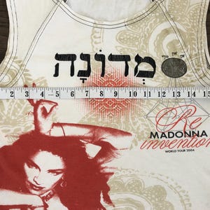 May include: Cream-colored tank top featuring a red and black graphic of Madonna. The top includes Hebrew text and the words "Re Invention World Tour 2004." The top measures approximately 16 inches across.