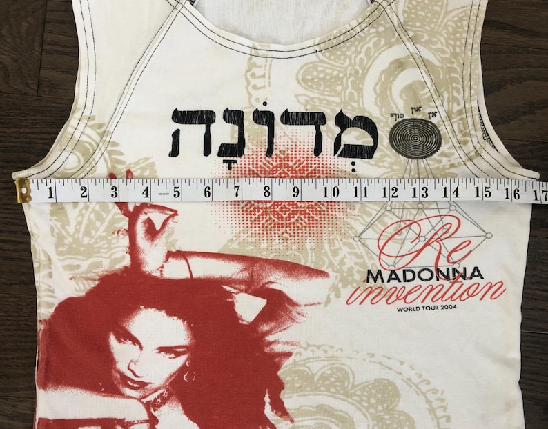 May include: Cream-colored tank top featuring a red and black graphic of Madonna. The top includes Hebrew text and the words "Re Invention World Tour 2004." The top measures approximately 16 inches across.