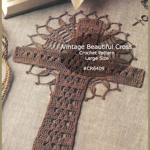 以下が含まれることがあります： 「Vintage Beautiful Cross Crochet Pattern Large Size #CR6409」の文字が入った茶色の手編み十字架。 質感のある糸で作られ、詳細な透かし彫りのデザインが施されています。 十字架は質感のある表面に置かれています。