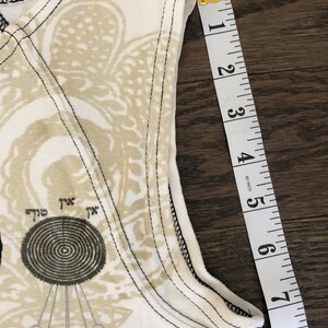May include: Close-up of a white tank top with a beige and black graphic design. The tank top is measured with a yellow and white measuring tape, showing approximately 8 inches.