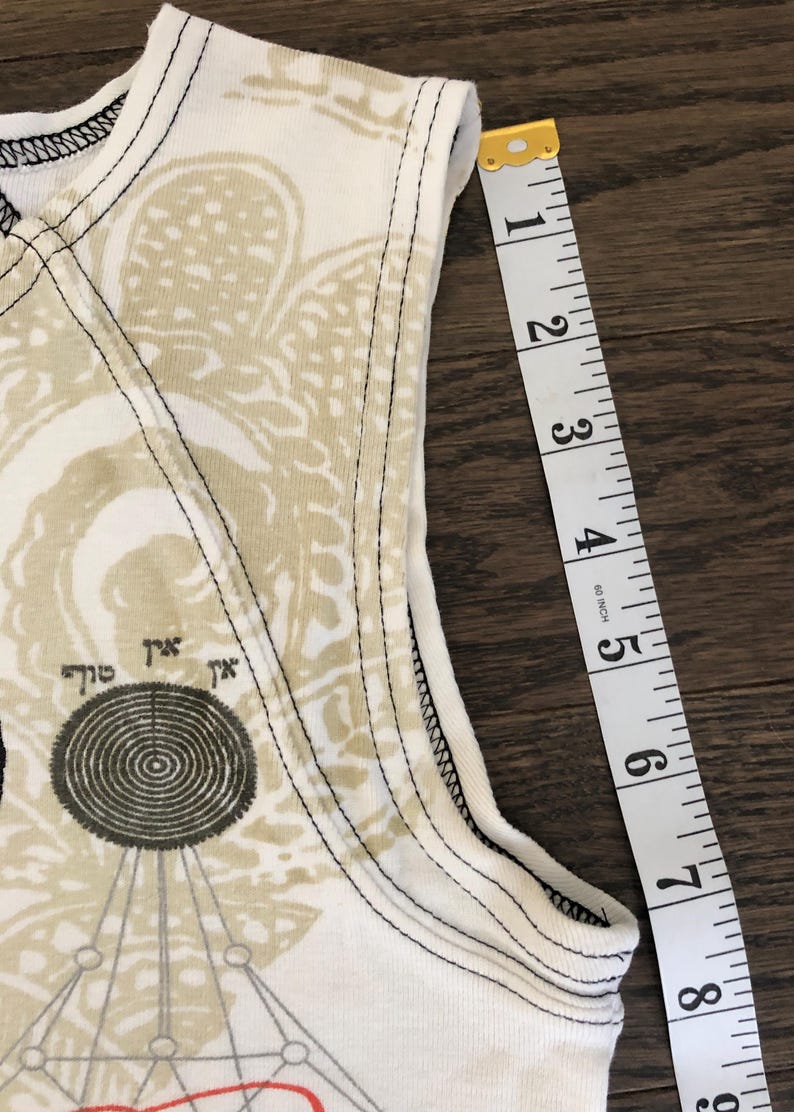 May include: Close-up of a white tank top with a beige and black graphic design. The tank top is measured with a yellow and white measuring tape, showing approximately 8 inches.