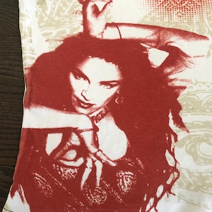 May include: White tank top featuring a red and white graphic of a woman with long hair and a hand gesture. The top has a beige pattern on the upper portion and black stitching along the bottom hem.