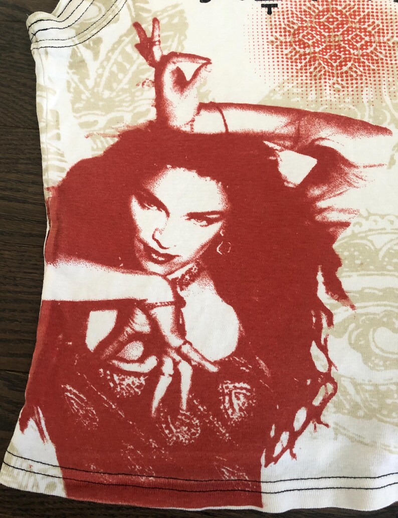 May include: White tank top featuring a red and white graphic of a woman with long hair and a hand gesture. The top has a beige pattern on the upper portion and black stitching along the bottom hem.
