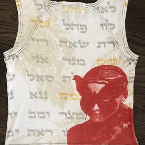 May include: White tank top with a red graphic of a person with a butterfly mask. The top features Hebrew text and a paisley design in the background. Black stitching outlines the neckline and armholes.