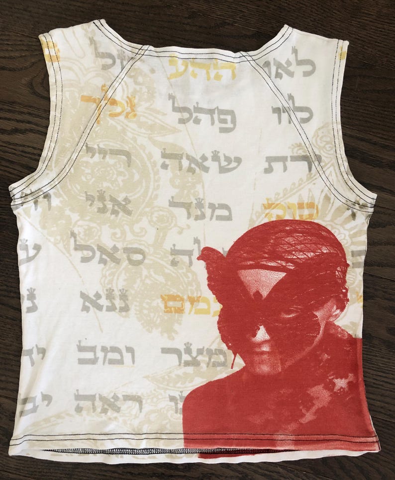May include: White tank top with a red graphic of a person with a butterfly mask. The top features Hebrew text and a paisley design in the background. Black stitching outlines the neckline and armholes.