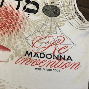 May include: White tank top featuring a red and beige design with the text "Re Madonna Invention World Tour 2004". The top has a geometric design and Hebrew text.