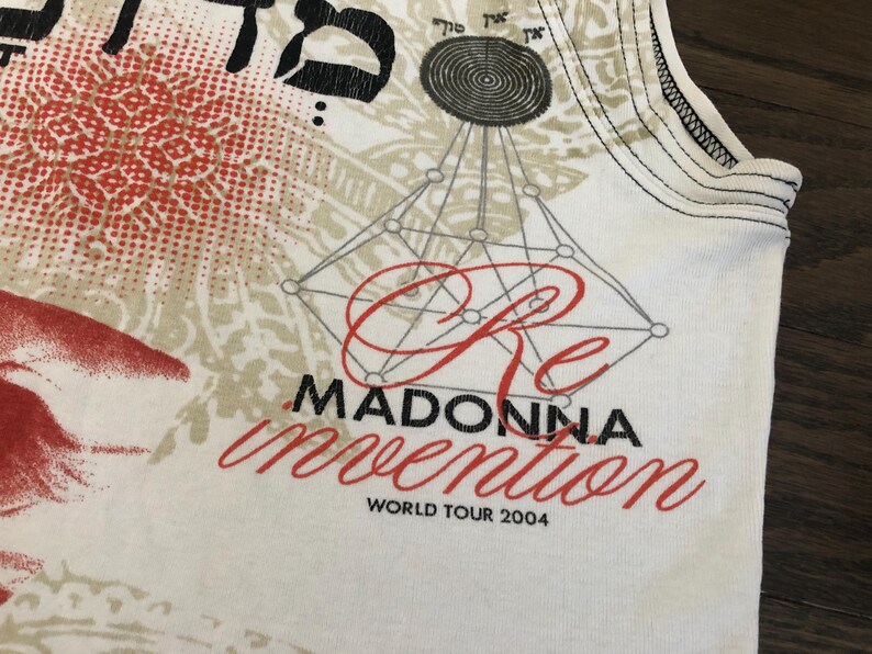 May include: White tank top featuring a red and beige design with the text "Re Madonna Invention World Tour 2004". The top has a geometric design and Hebrew text.