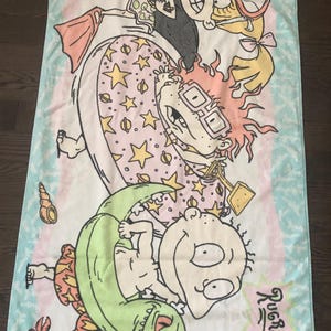 May include: A pink and blue beach towel featuring the Rugrats characters. Tommy Pickles is riding a green dinosaur float, Chuckie Finster is in a green inflatable ring, Phil and Lil DeVille are in a pink and yellow inflatable ring, and Angelica Pickles is holding a red and white life preserver.