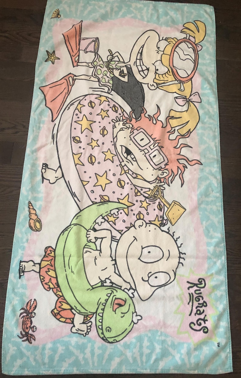 May include: A pink and blue beach towel featuring the Rugrats characters. Tommy Pickles is riding a green dinosaur float, Chuckie Finster is in a green inflatable ring, Phil and Lil DeVille are in a pink and yellow inflatable ring, and Angelica Pickles is holding a red and white life preserver.