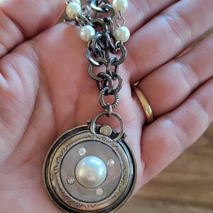 May include: A silver chain necklace with a round pendant featuring a large pearl surrounded by smaller rhinestones. The chain is adorned with white pearl beads.