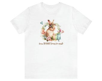 May include: White t-shirt with a watercolor illustration of a brown bunny wearing a flower crown and holding a stack of books. The text "Some BUNNY loves to read!" is printed below the bunny.