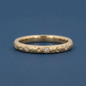 May include: A gold ring with a celestial design. The ring features stars, moons, and small diamonds. The ring is set against a dark blue background.