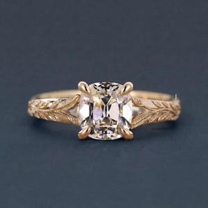 Op de afbeelding: Een gouden ring met een centrale edelsteen in kussenslijpvorm, vastgehouden door vier klauwen. De ringband is voorzien van een ingewikkeld, gegraveerd bladpatroon dat zich vanaf de zetting langs beide zijden van de schacht uitstrekt.