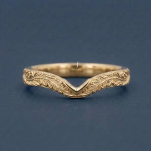 May include: A gold-colored, V-shaped ring with an intricate leaf pattern. The ring is set against a dark blue background. The ring's design features a delicate, engraved floral motif.