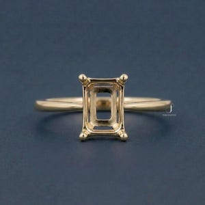 May include: A gold ring setting with an emerald cut design. The ring features a rectangular centre setting with four prongs and a delicate band. The ring is set against a dark blue background.