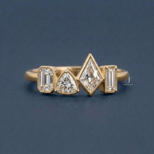 May include: A gold ring featuring four clear, geometric-cut gemstones: an emerald cut, a triangle cut, a kite cut, and another emerald cut. The ring has a delicate band and is set against a dark blue background.