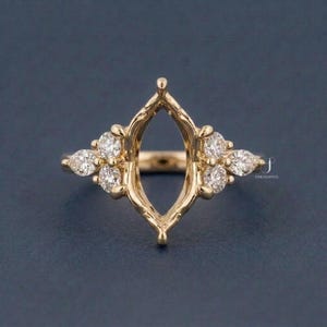 May include: A gold ring setting with a central oval opening, surrounded by small, sparkling diamonds. The ring has a unique, ornate design with diamond clusters on either side, set against a dark blue background.