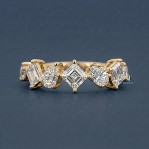May include: A gold band ring featuring a series of differently shaped clear gemstones. The gemstones include emerald cut, pear cut, oval cut, and square cut stones. The ring is set against a dark blue background.