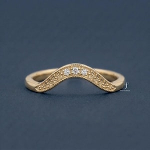May include: A gold-colored curved ring with three small, clear gemstones set in the center. The ring features a detailed leaf pattern along the curved top. The ring is set against a dark blue background.