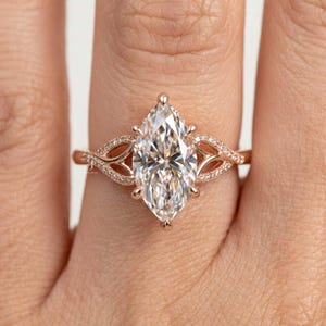 May include: A rose gold ring featuring a large marquise-cut centre stone. The band is crafted with intricate, openwork metal detailing on both sides, accented with small, round, pavé-set stones that extend along the shoulders of the ring.